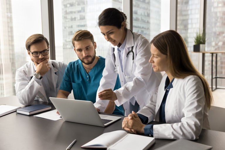 Young Indian medical team leader presenting job application on laptop to colleagues. Diverse doctors meeting at computer, talking on group video call, looking at screen, speaking, listening